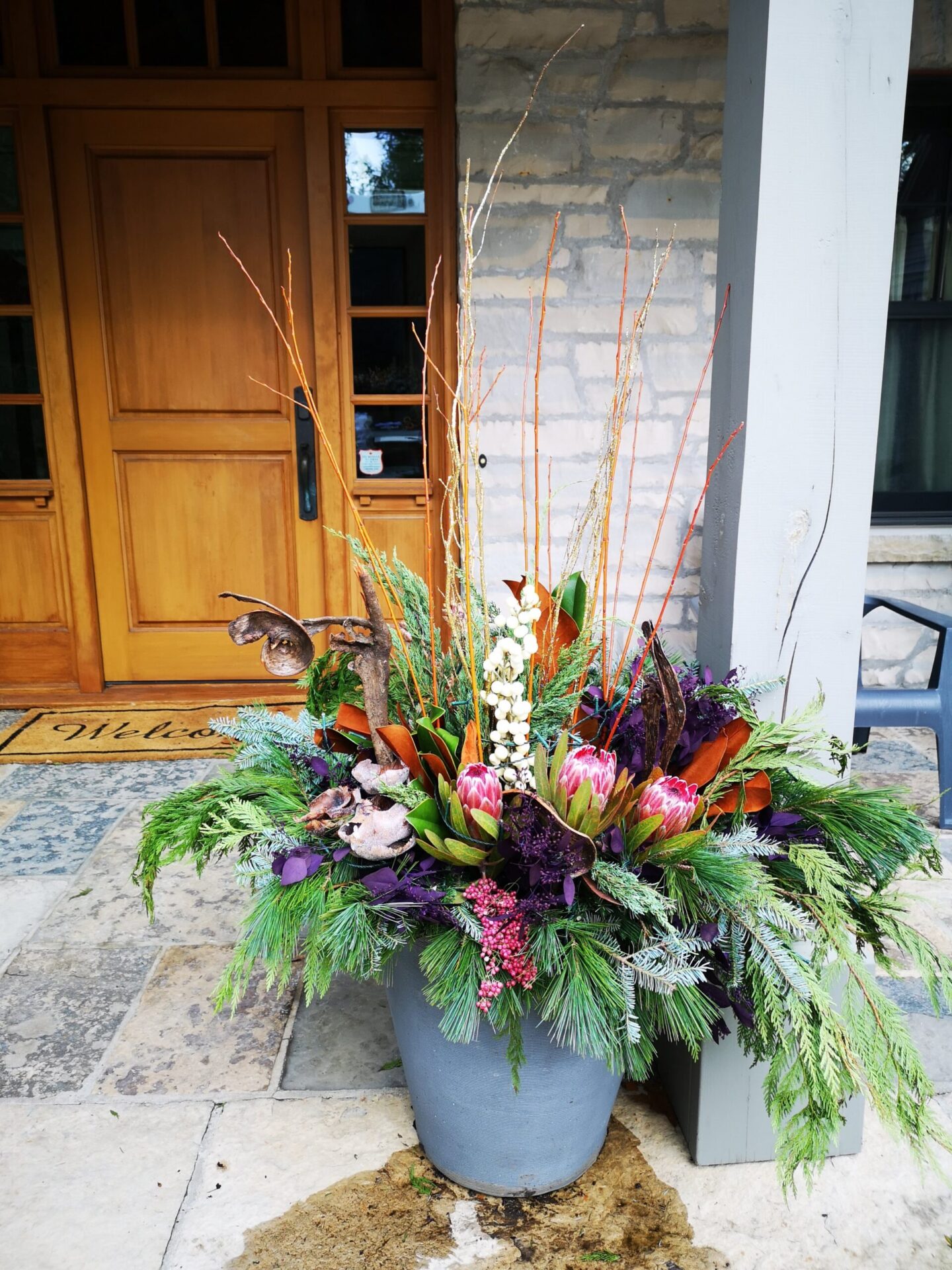 This image shows a colorful floral arrangement in a gray pot at a doorway, featuring greenery, berries, and twigs, creating a welcoming atmosphere.