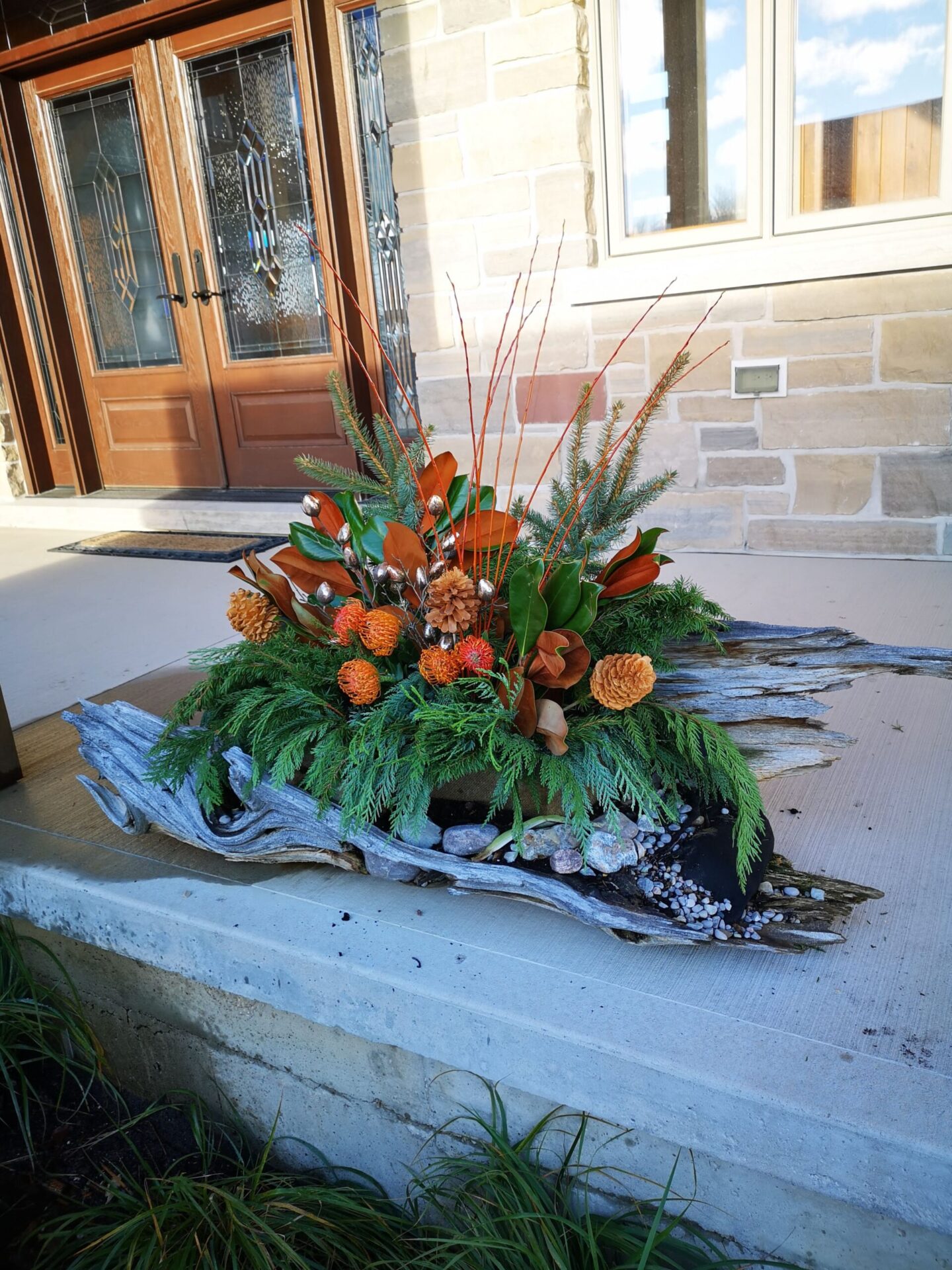 A festive floral arrangement on weathered wood, comprising orange blooms, pine cones, and evergreen foliage, placed on a residential entryway ledge.