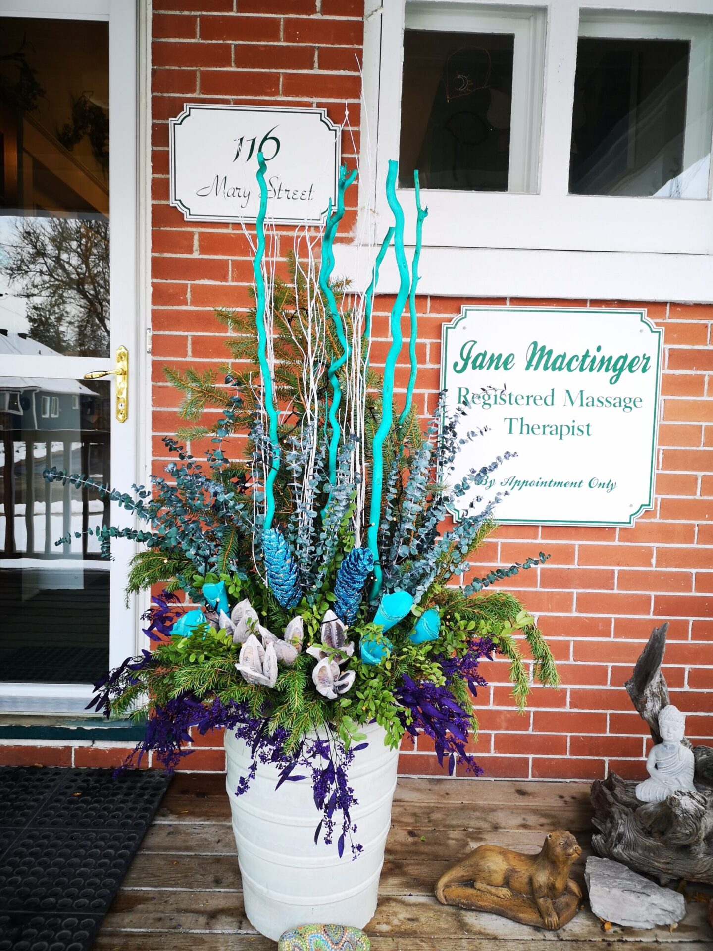 An ornate floral arrangement in a white pot, featuring blue accents, sits on a porch by a door with informational signs, on a wooden deck.