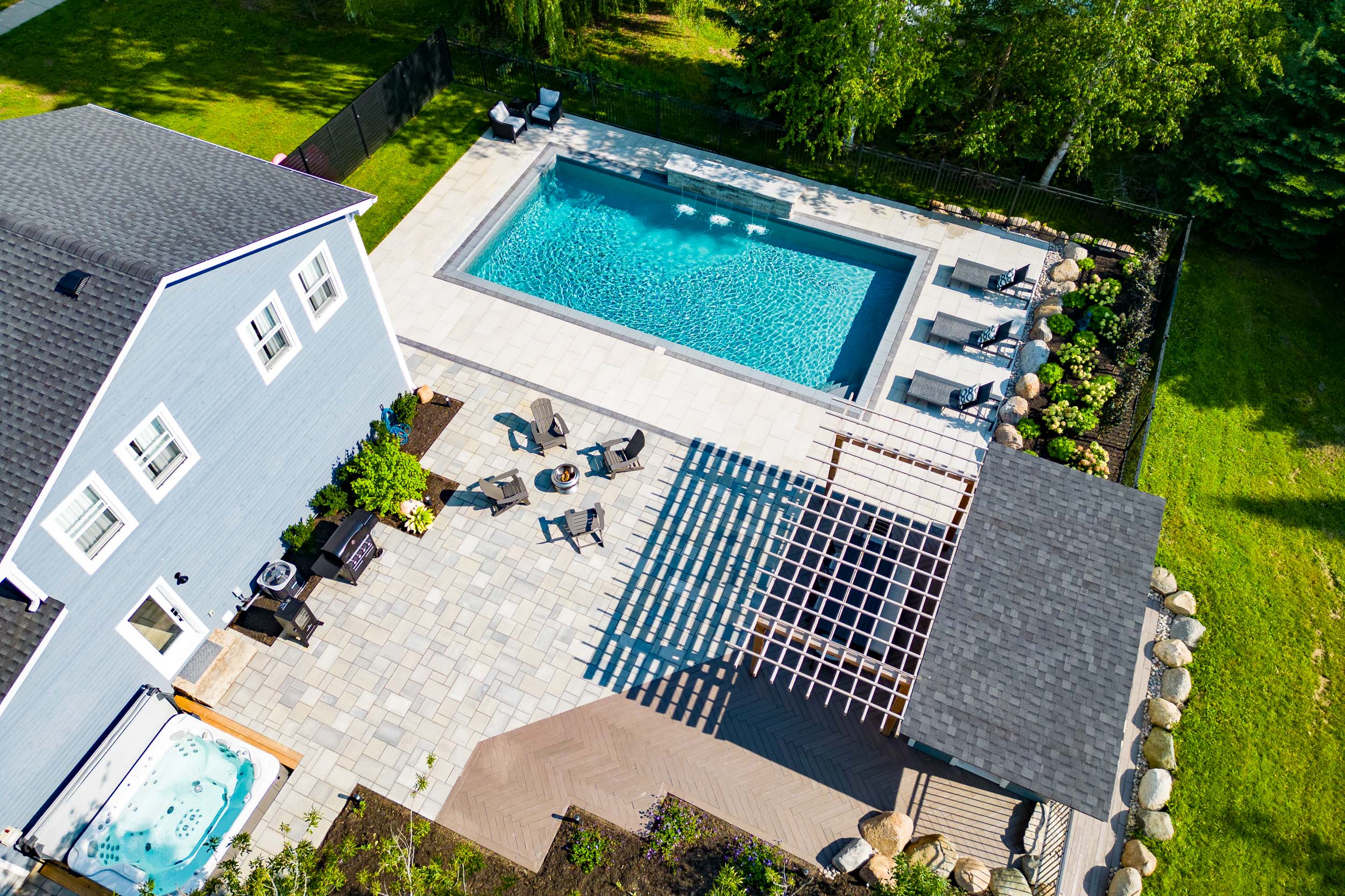 An aerial view of a residential backyard with a swimming pool, hot tub, patio, outdoor furniture, and a manicured lawn surrounded by a fence.