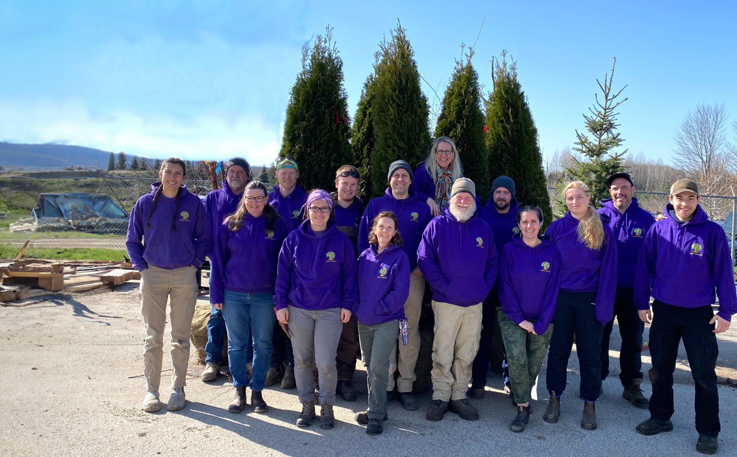 A group of people in matching purple jackets smiling outdoors, with tall conifer trees and a clear blue sky in the background.