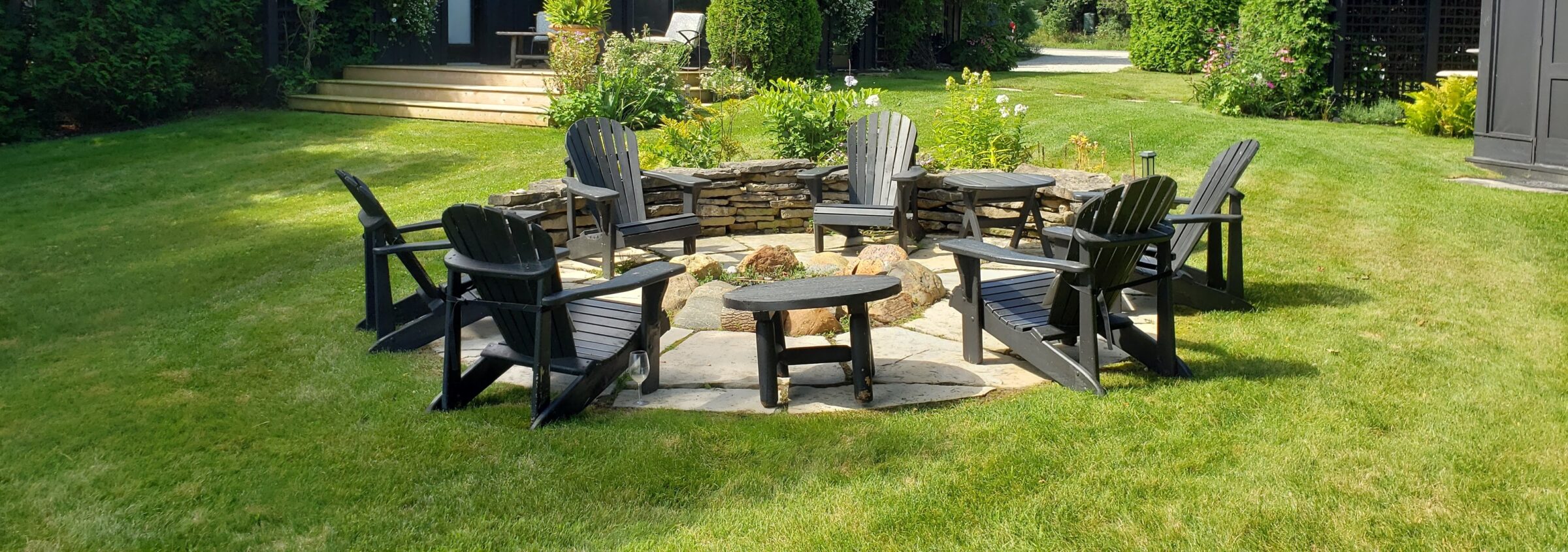 A serene garden with green grass features Adirondack chairs surrounding a fire pit; stepping stones, landscaped plants, and a stone wall enhance the setting.