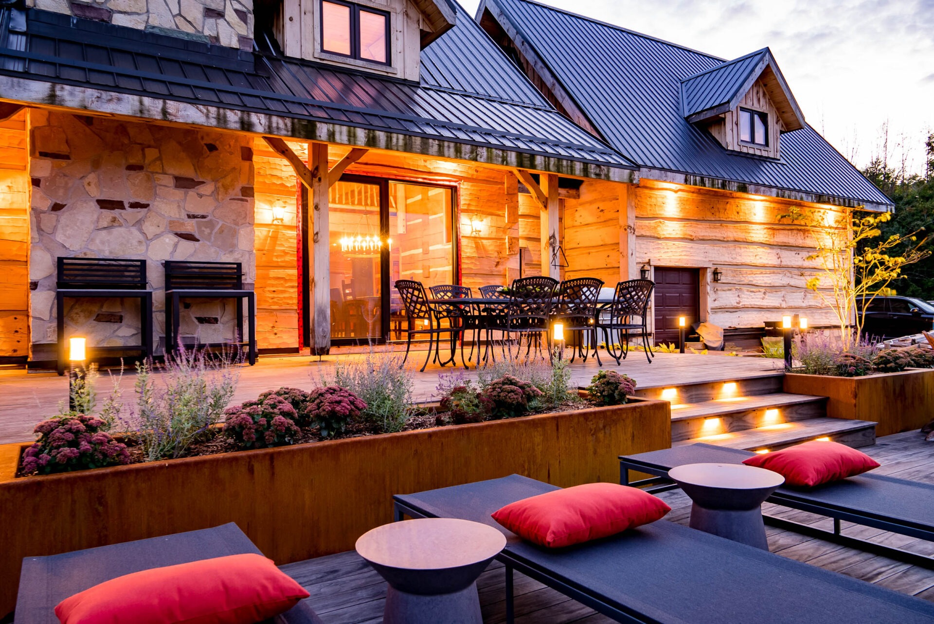 A cozy outdoor patio of a log cabin with warm lighting, comfortable seating, a dining area, and lush plants during twilight.