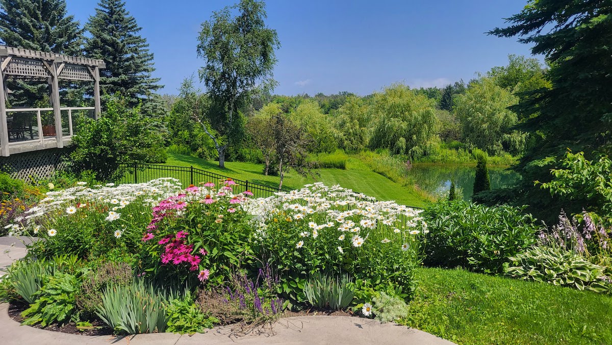 A tranquil garden with colorful flowers, a wooden gazebo, lush greenery, and a pond reflecting trees under a clear blue sky on a sunny day.