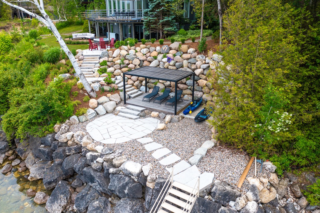 An aerial view of a landscaped waterfront property with a gazebo, loungers, steps leading to a house, and a rocky shore.