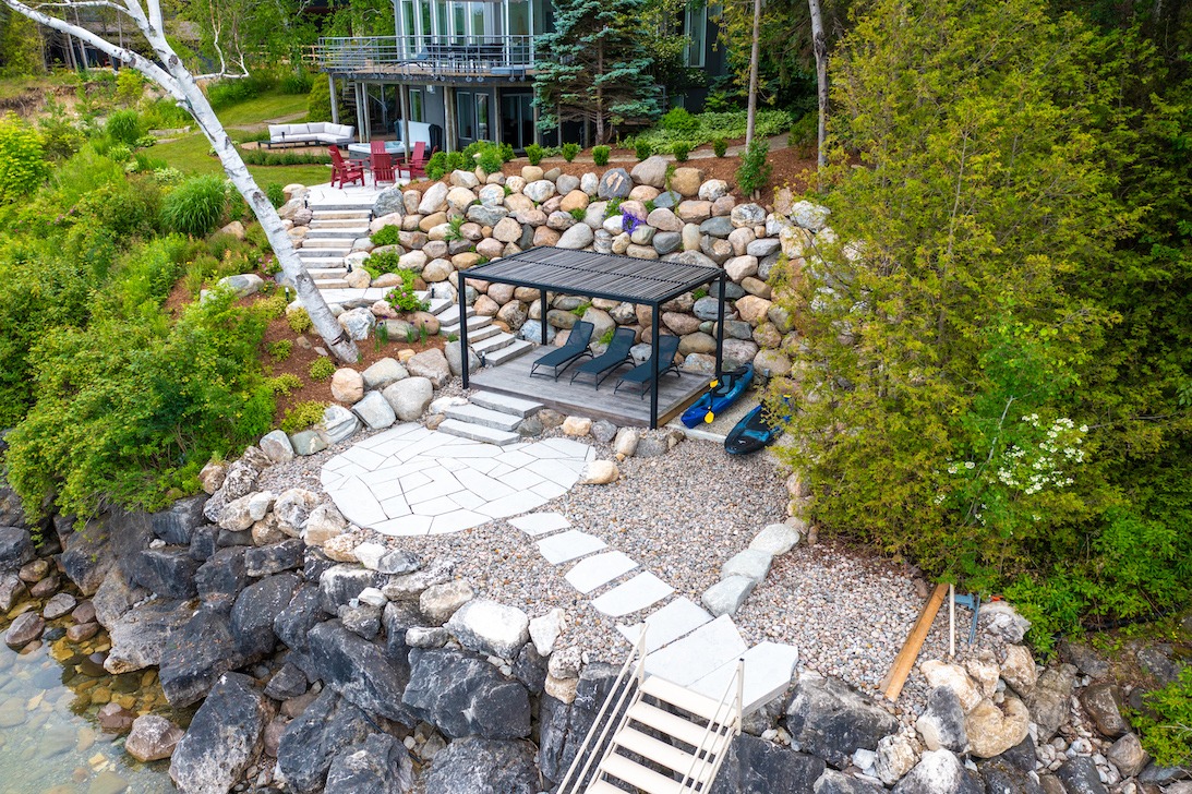 Aerial view of a landscaped backyard with a gazebo, stone steps, and pebble pathways along a rocky waterfront, adjacent to a modern house.