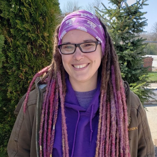 A smiling person with glasses and purple headband stands outdoors. They have long, burgundy and blonde dreadlocks, a nose ring, and are wearing a purple hoodie.