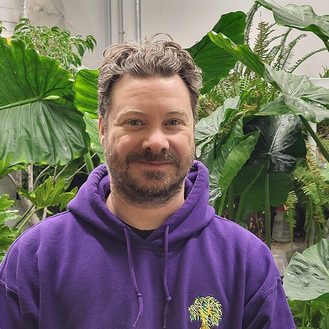A person with short curly hair and facial stubble is wearing a purple hoodie, smiling gently in a greenhouse surrounded by large green plants.