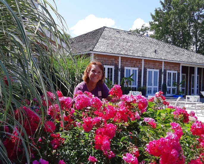 A smiling person is peeking through vibrant pink flowers in a sunny garden with a traditional house and lush greenery in the background.