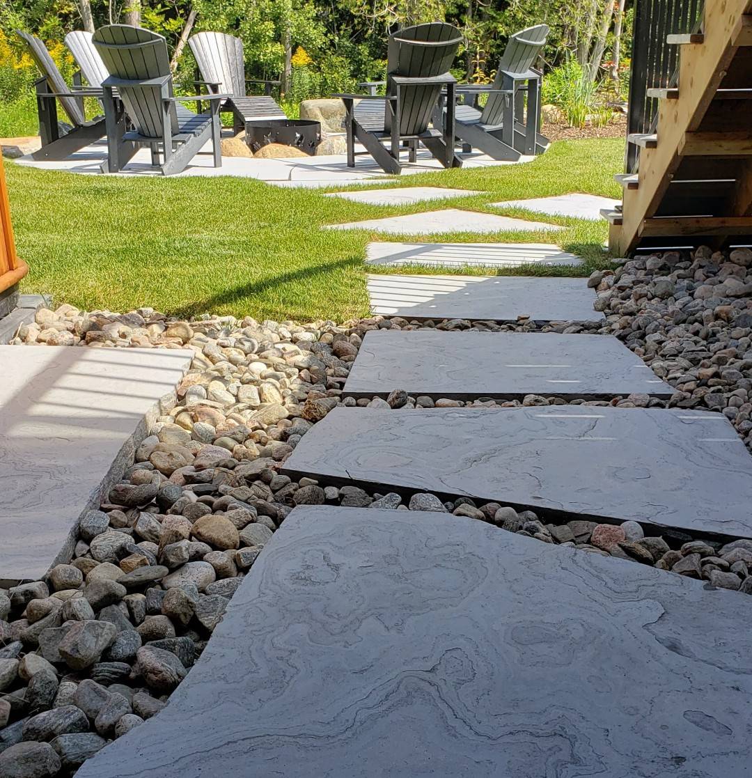 Stepping stones form a pathway through a landscaped yard with river rocks to a wooden deck with Adirondack chairs and a fire pit, suggesting a cozy outdoor space.