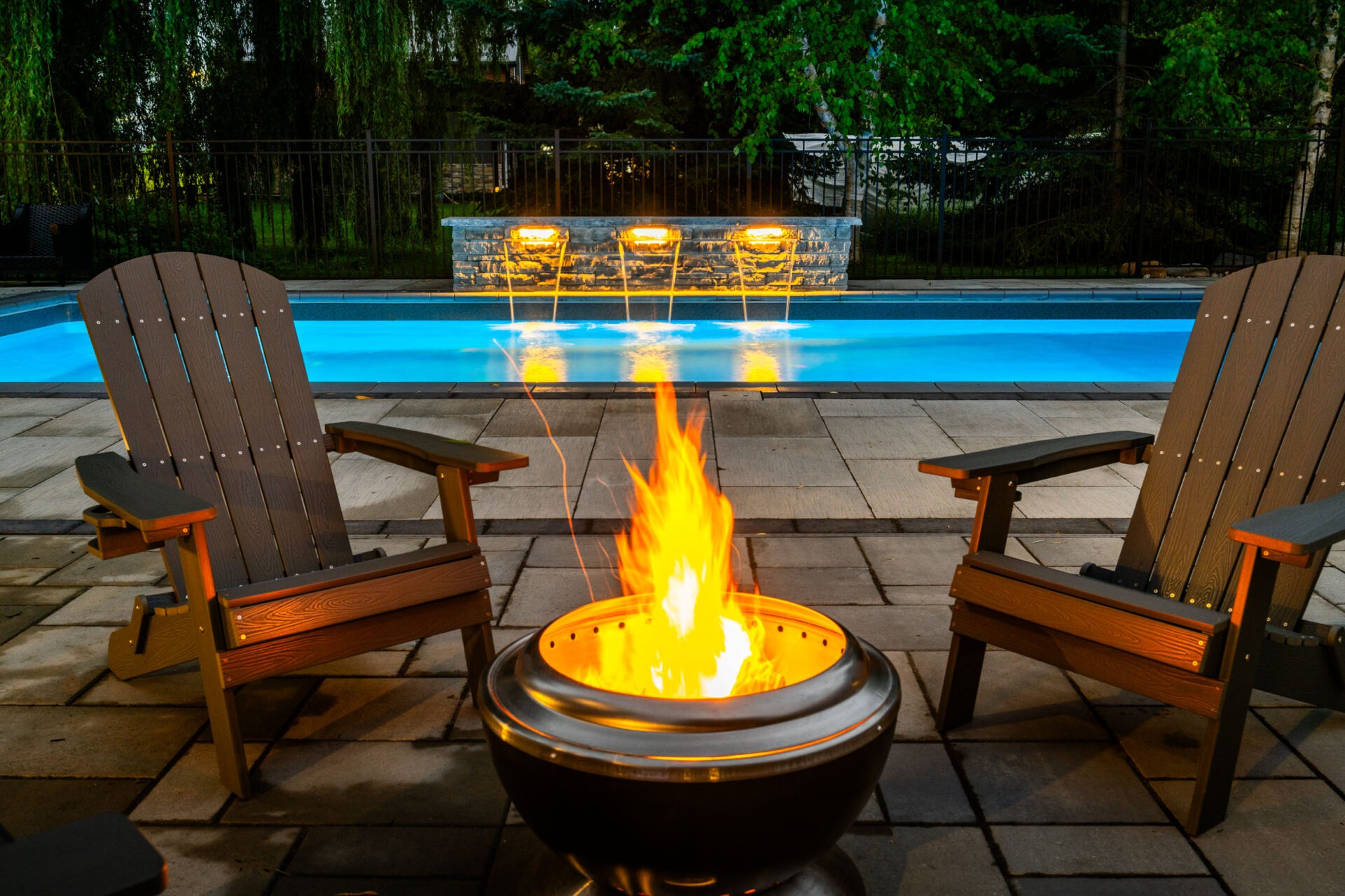 Two wooden Adirondack chairs flank a blazing fire pit on a patio, with a lit pool and stone fire feature in the background at dusk.