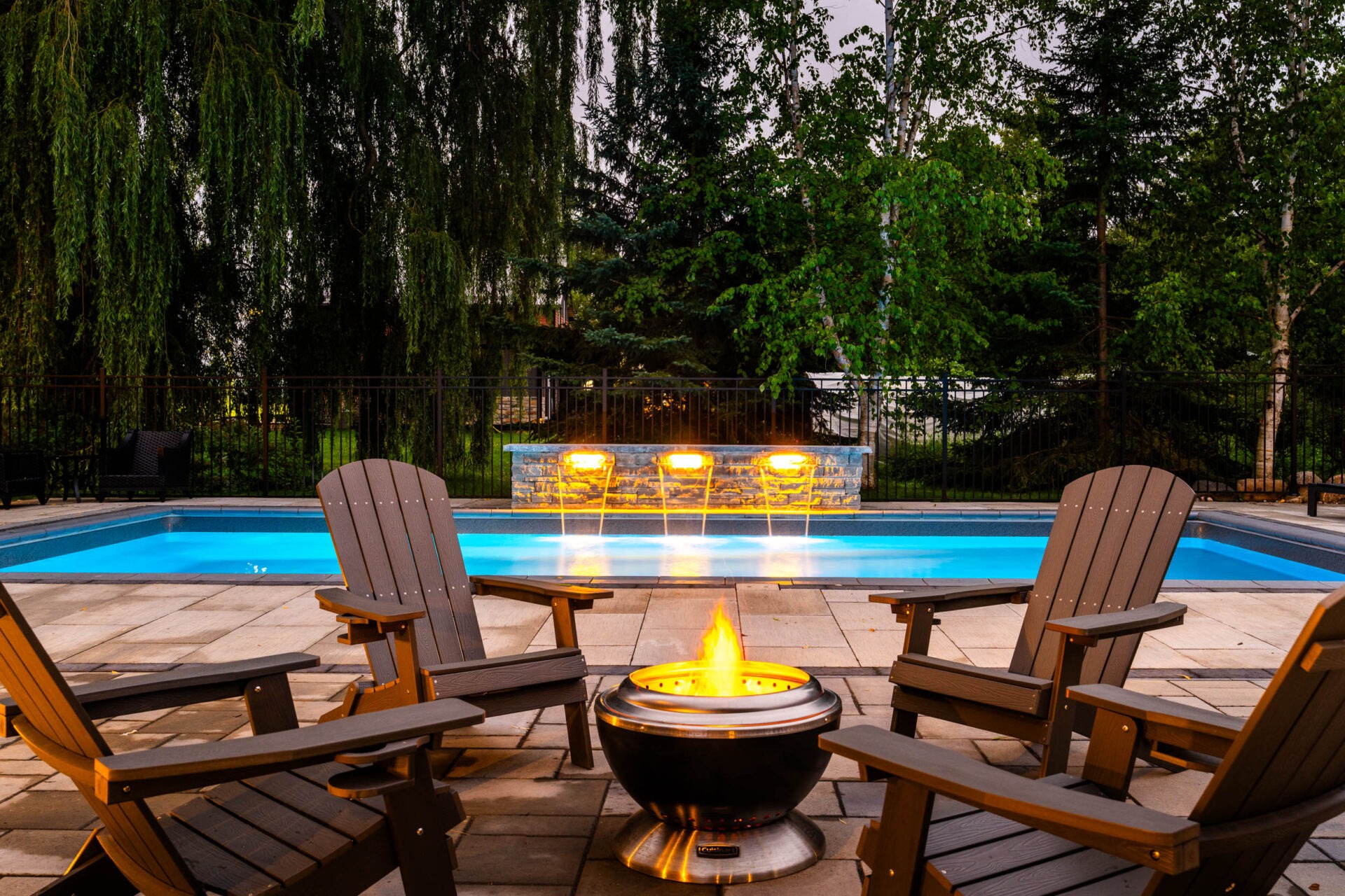 An outdoor setting at twilight with a lit fire pit, Adirondack chairs, a swimming pool and trees in the background, fenced for privacy.