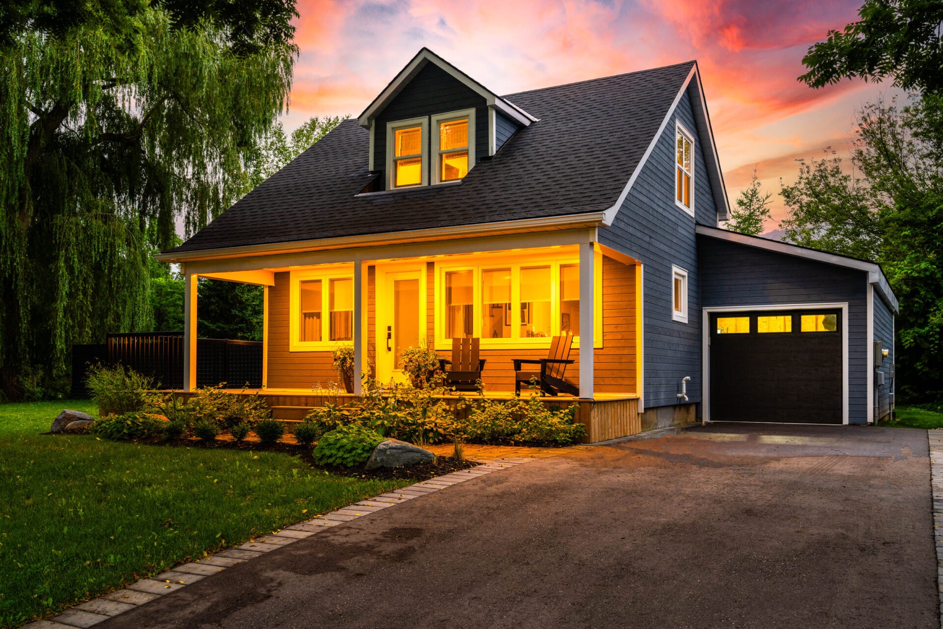 Charming two-story house with gray siding and yellow-lit windows during twilight. Cozy porch with chairs, attached garage, lush greenery, and a vibrant sky.