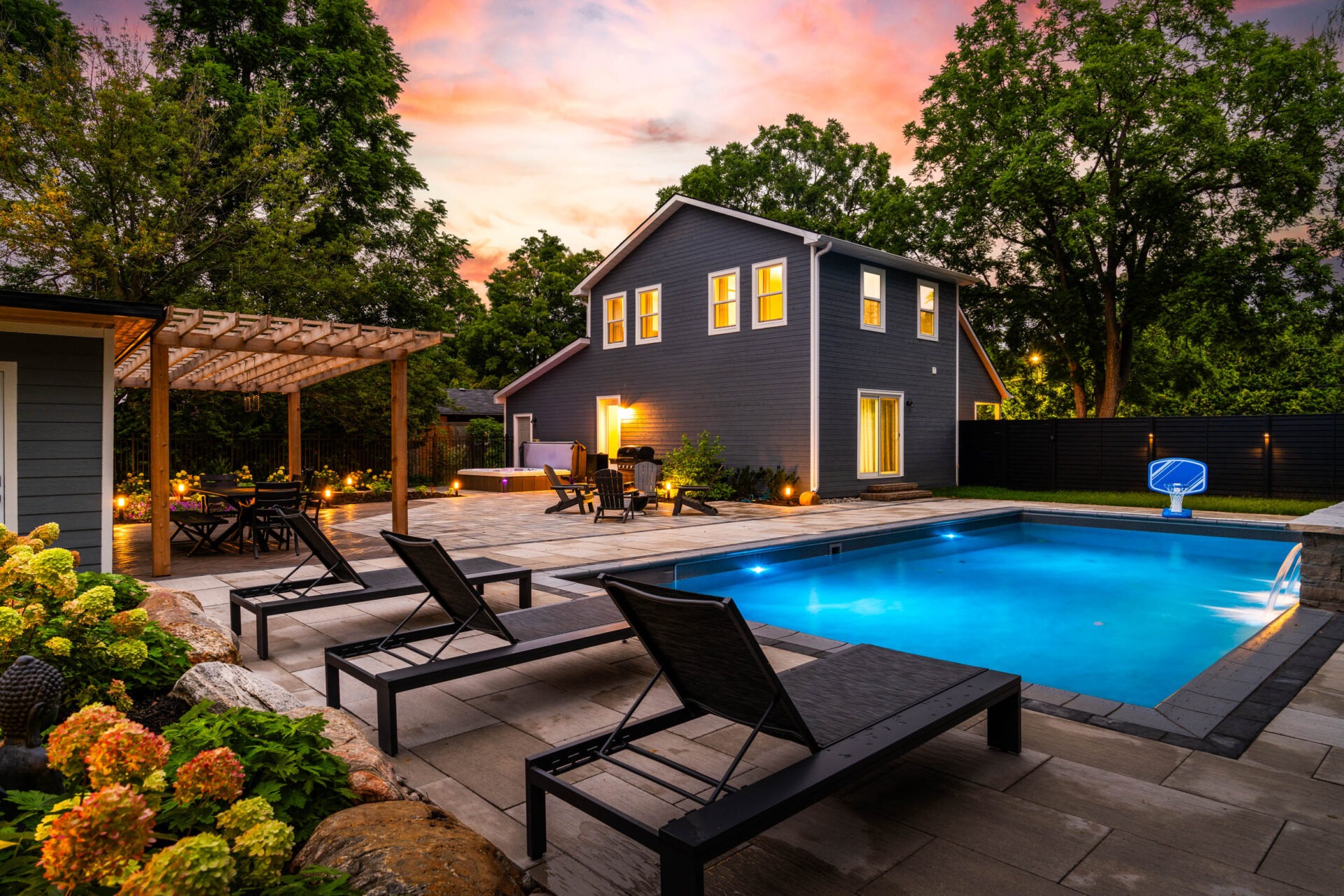 A backyard at dusk featuring a modern two-story house with a swimming pool, lounge chairs, pergola dining area, and ambient lighting under a sunset sky.