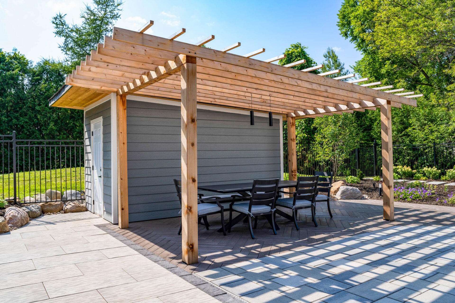A wooden pergola with a partial lattice design extends over a paved patio with a dining set beside a grey shed, set against a landscaped garden and clear sky.
