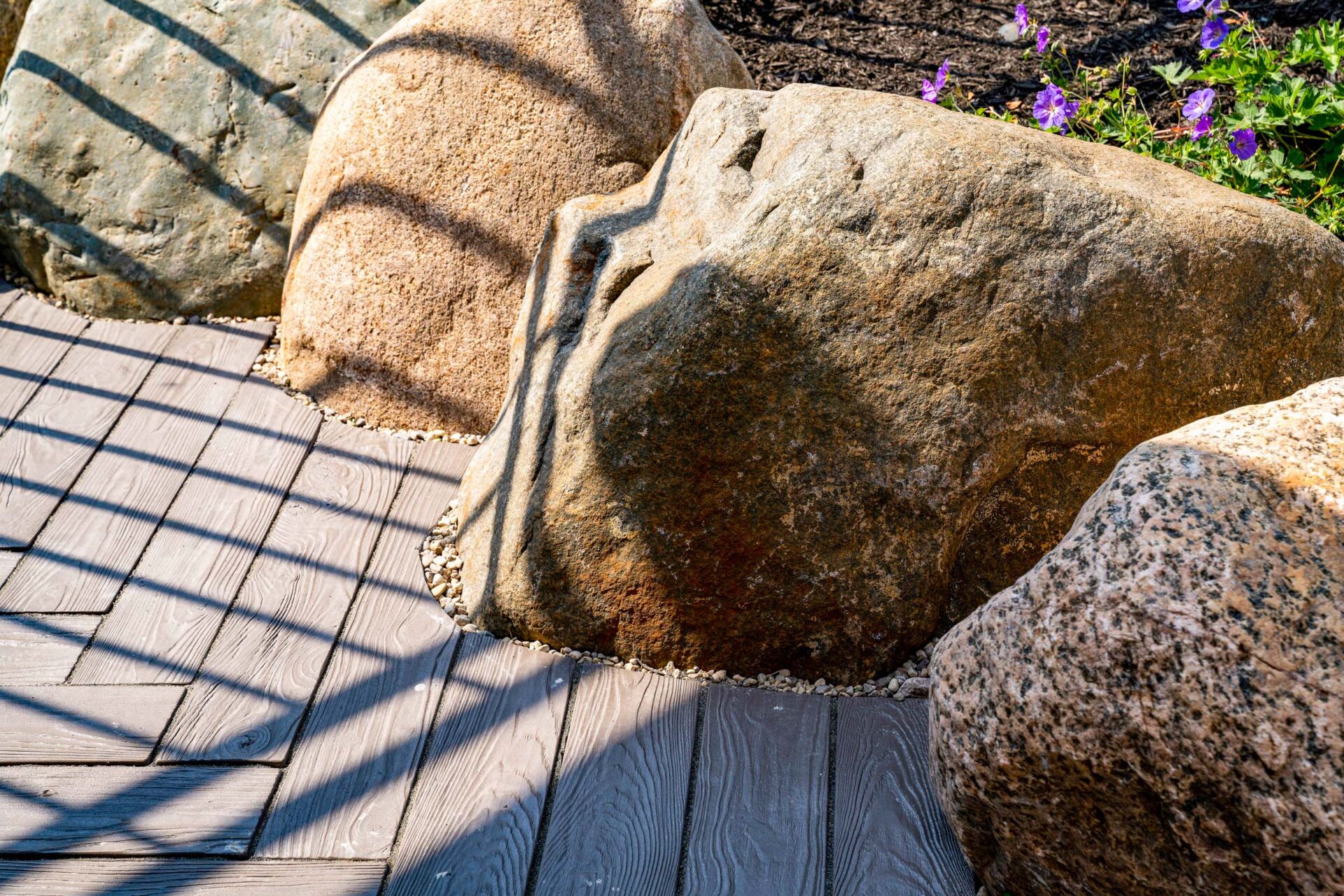 The image shows large rocks positioned beside a wooden boardwalk with shadows creating patterns, complemented by purple flowers in the background.