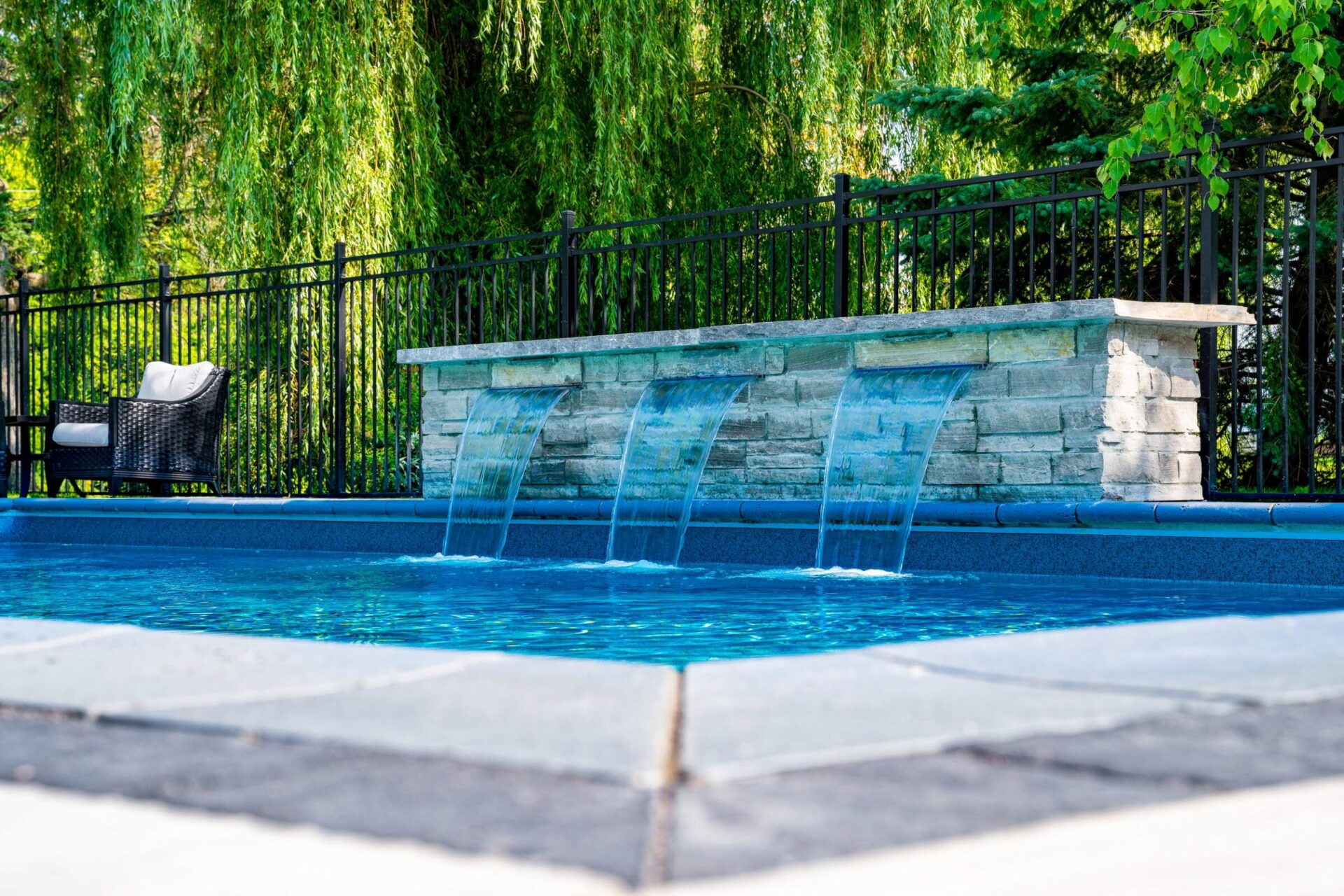 An inviting outdoor swimming pool with a cascading stone waterfall feature, surrounded by lush greenery and a single woven chair beside a metal fence.