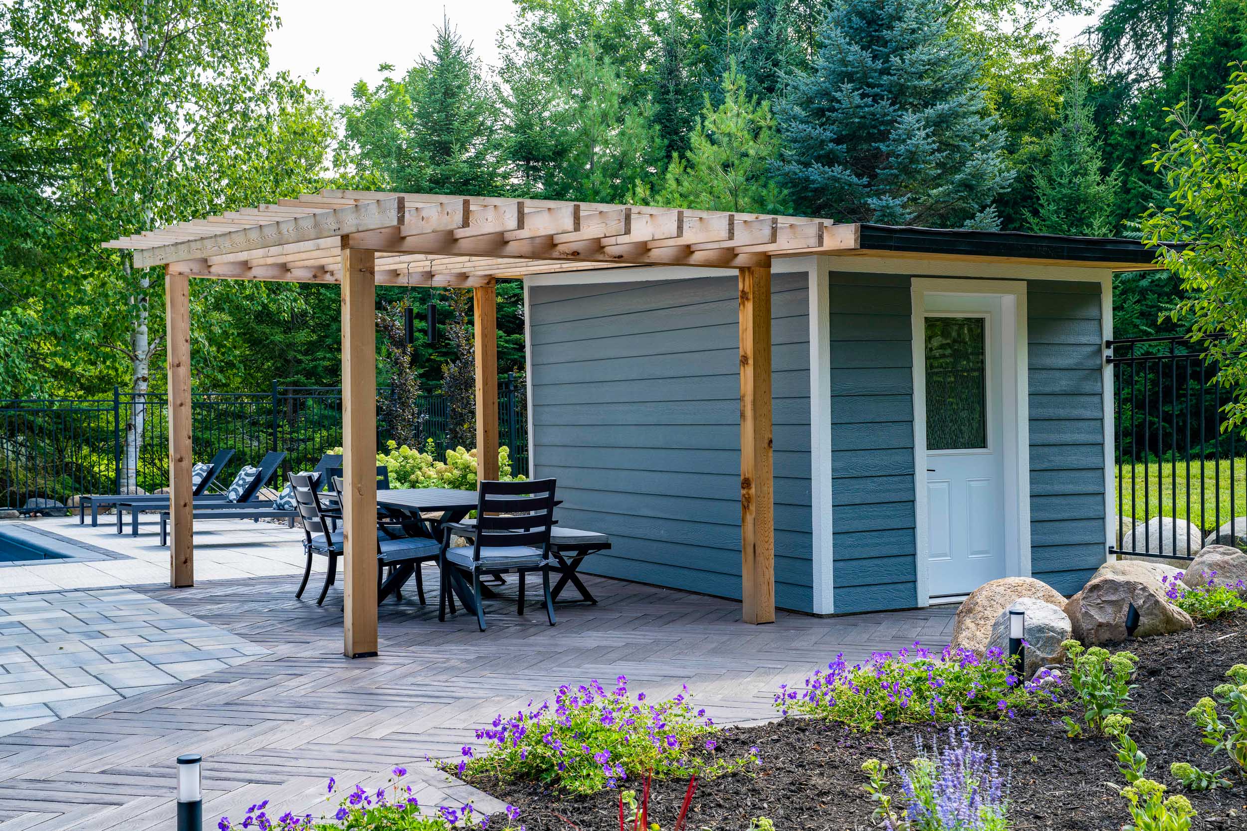 A modern backyard with a wooden pergola, a small shed, an outdoor dining set, landscaped garden, and a fenced pool area with lounge chairs.