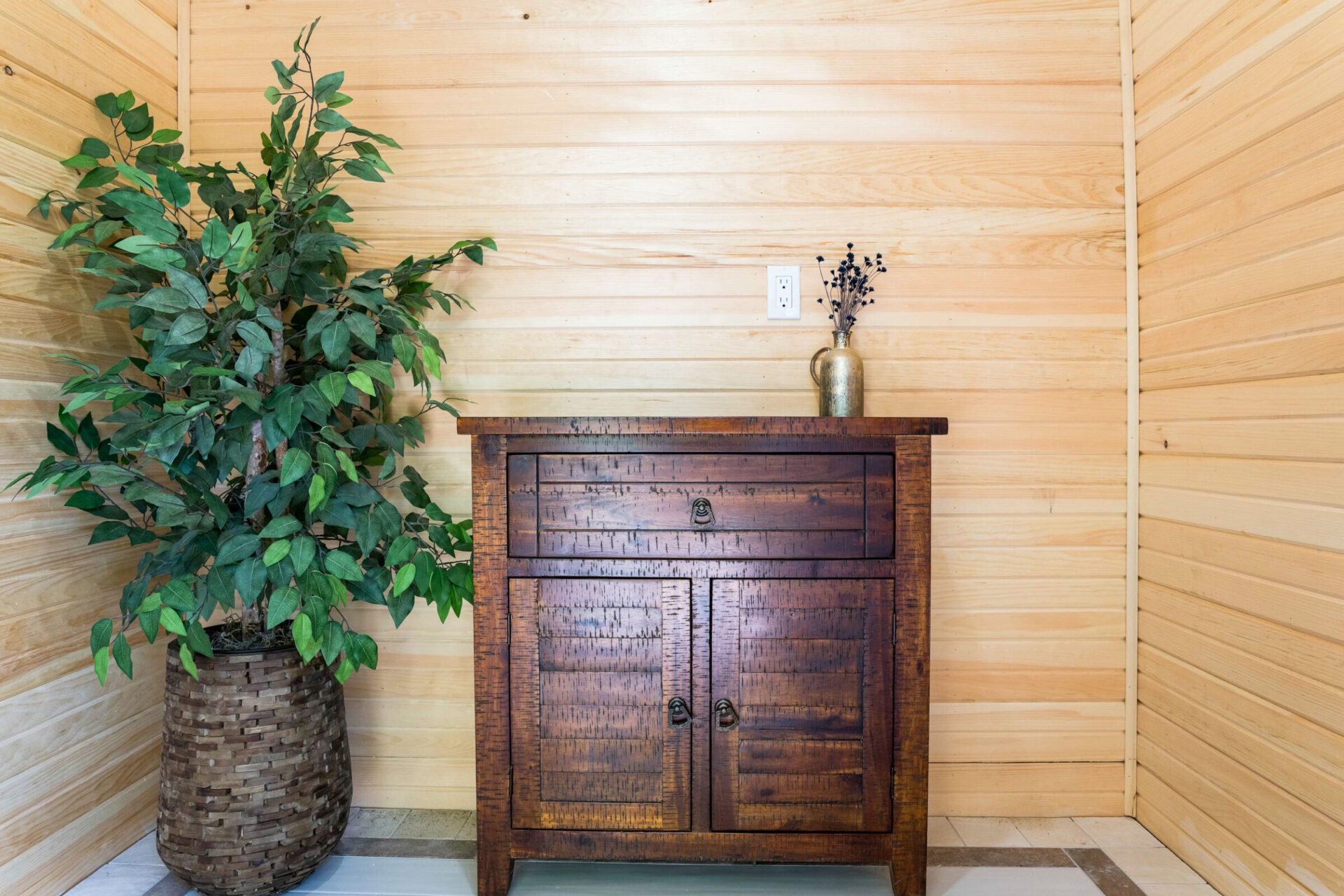 A room corner featuring a large indoor plant in a woven basket and a rustic wooden cabinet with a vase of dried flowers atop it, against a wood-paneled wall.