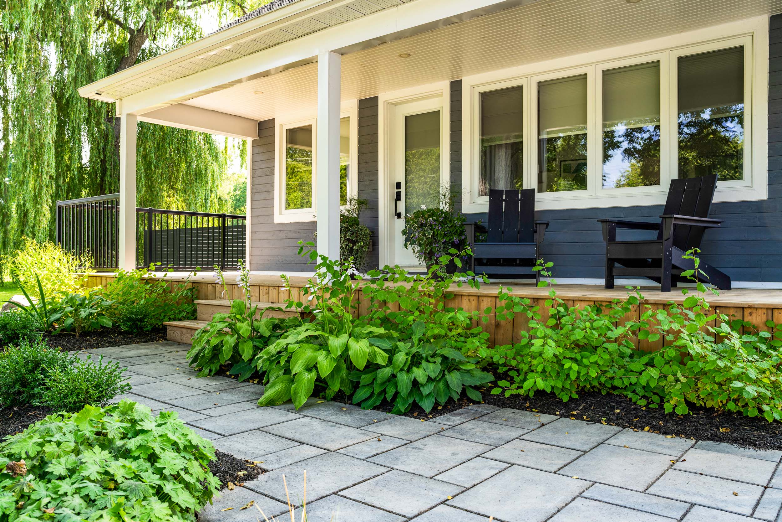 A cozy house porch with two black Adirondack chairs, surrounded by lush greenery and a well-maintained garden, under a bright, clear sky.