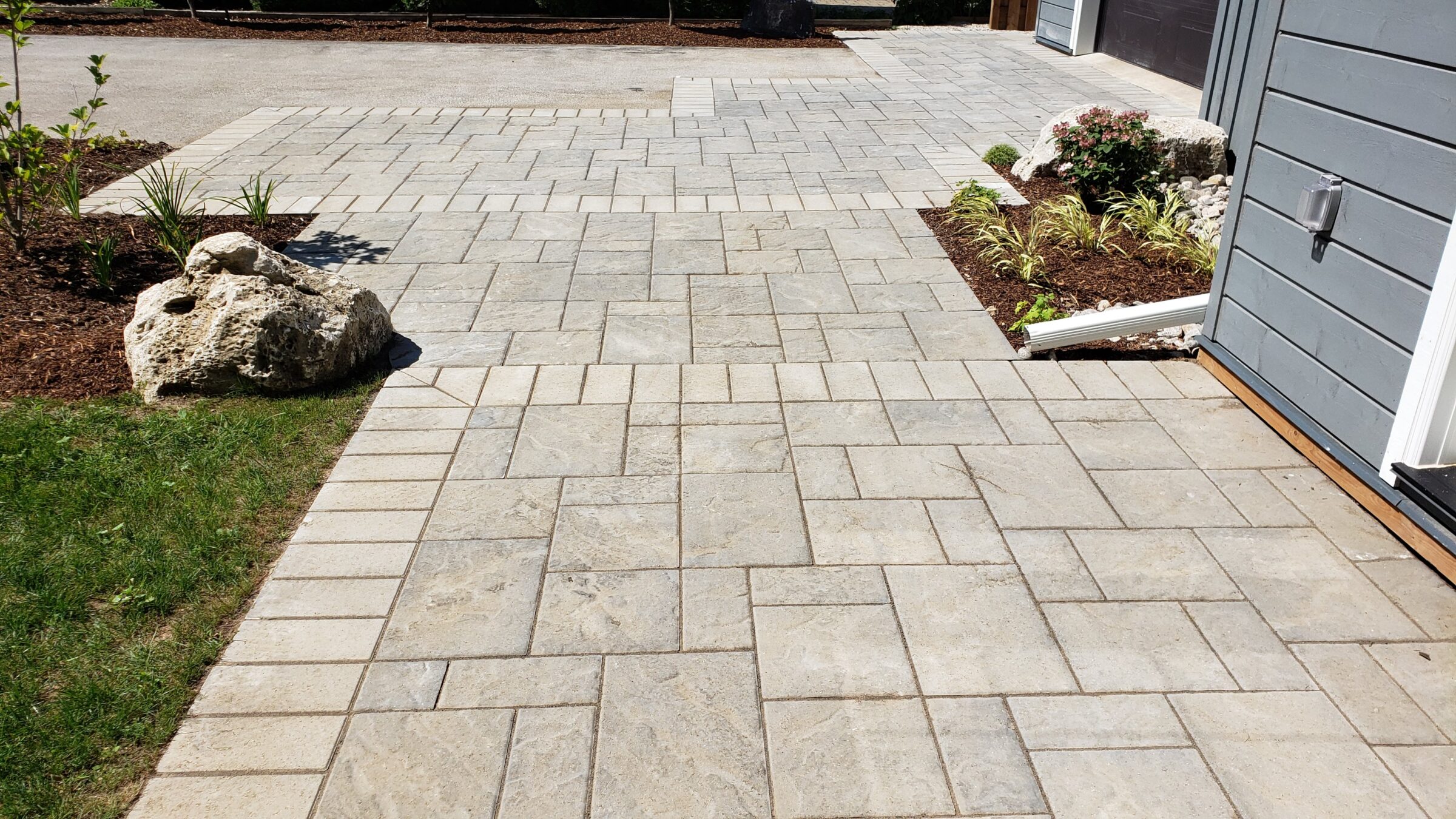 A well-organized backyard with patterned paving stones leading to an outbuilding. Landscaping with mulch, plants, and decorative rocks completes the serene scene.