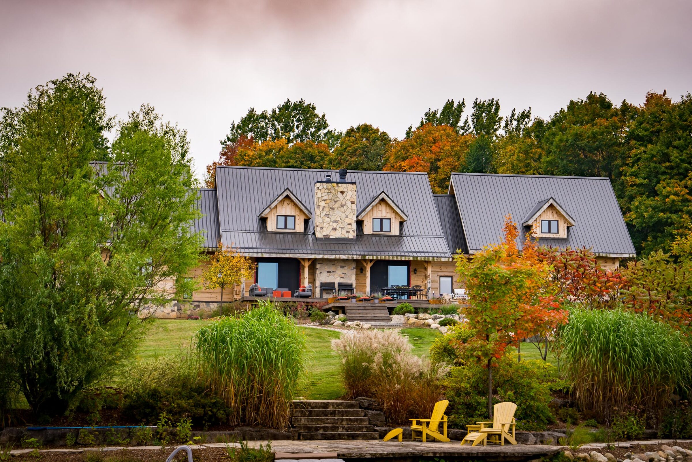 Stone and wood house with metal roofs, nestled in autumn foliage. Landscaped garden with ornamental grasses, steps, and bright yellow Adirondack chairs.