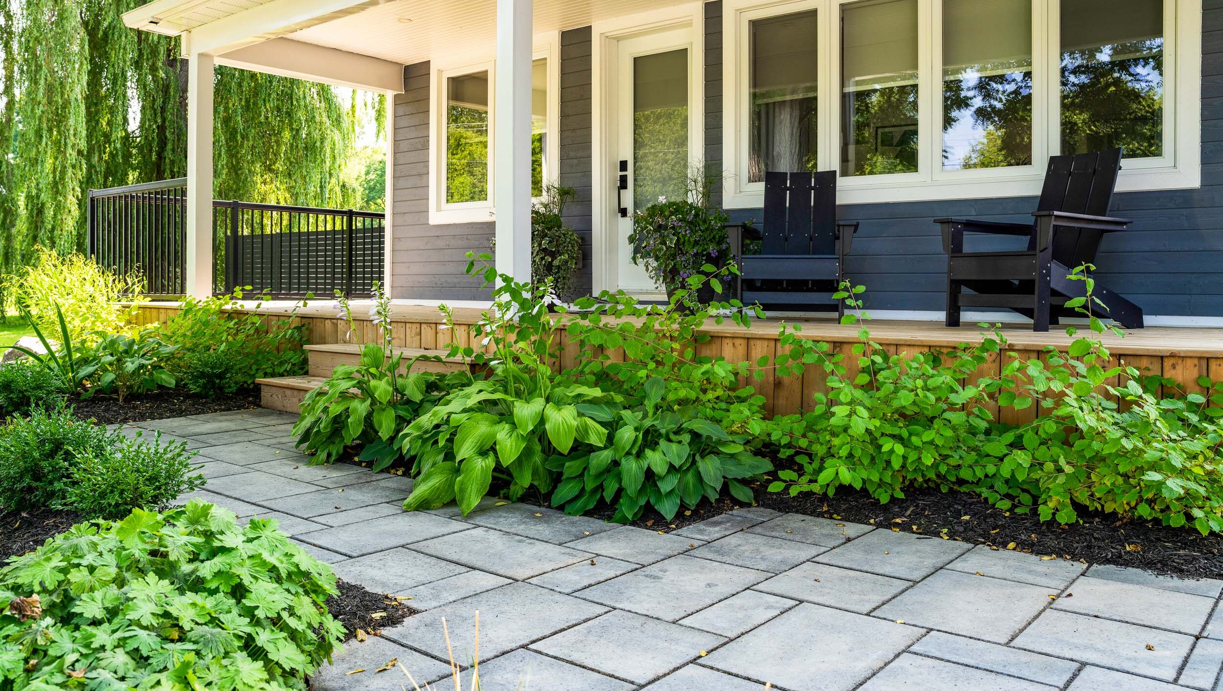 This image shows a cozy home porch with two black Adirondack chairs, lush greenery, stone pathways, and manicured landscaping under a clear sky.