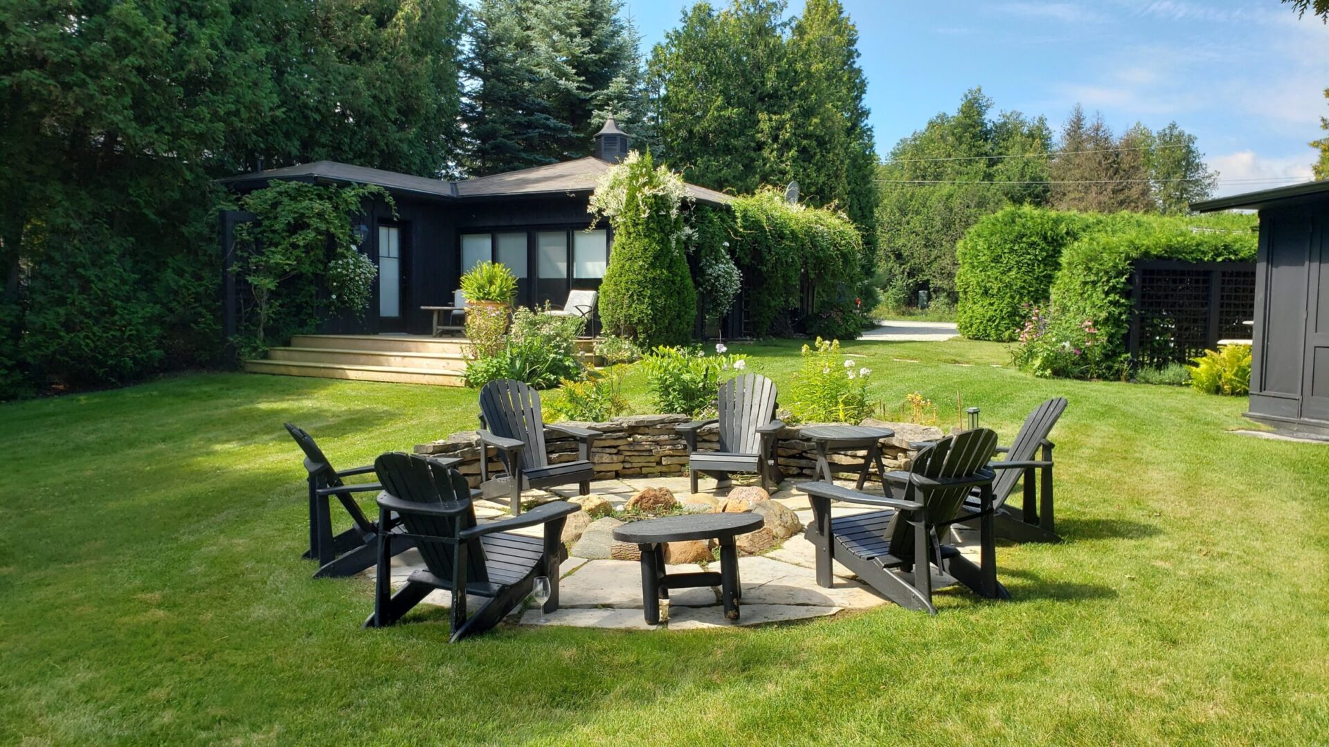 This image shows a cozy backyard with a fire pit surrounded by Adirondack chairs. A lush lawn, garden beds, hedges, and a black modern house are visible.