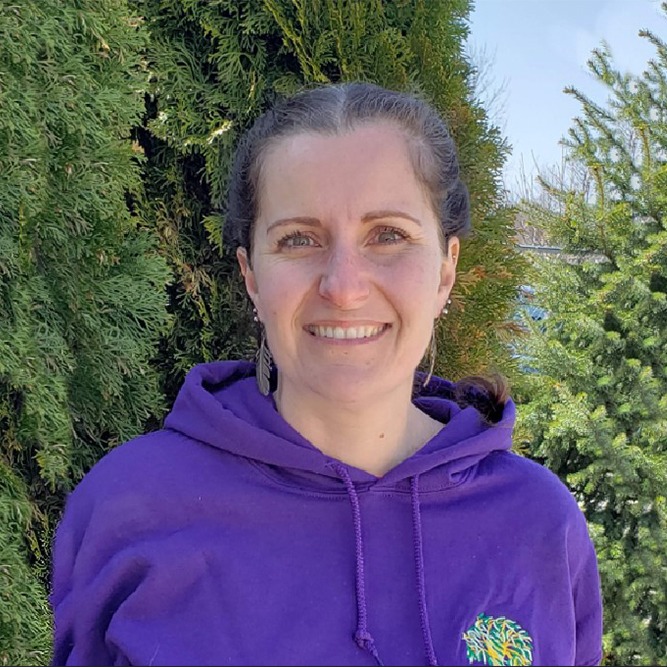 A person in a purple hoodie is smiling in front of a green leafy backdrop on a sunny day, with a casual and friendly demeanor.
