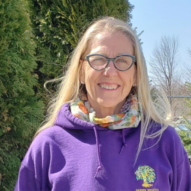 A smiling person with long blonde hair and glasses stands outdoors. They are wearing a purple hoodie and a colorful scarf, with trees in the background.