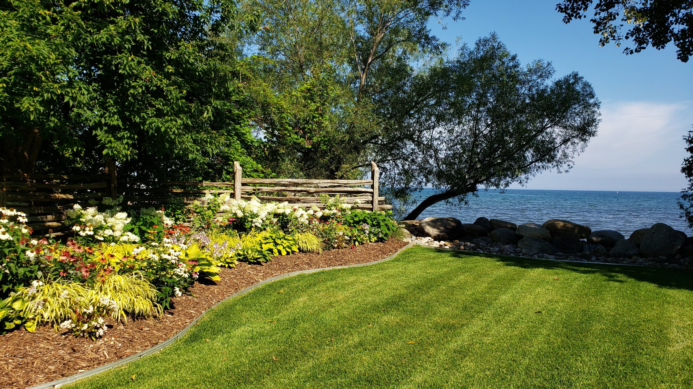 A serene lakeside garden with a wooden fence, lush flowerbeds, neatly maintained grass, large rocks, and trees under a clear blue sky.