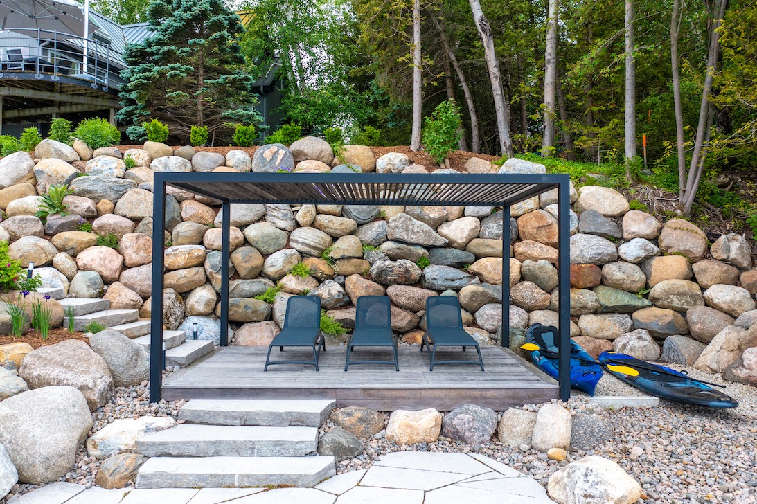 A modern outdoor seating area with a pergola, two chairs, and a kayak beside it, set against a backdrop of large, colorful boulders and greenery.