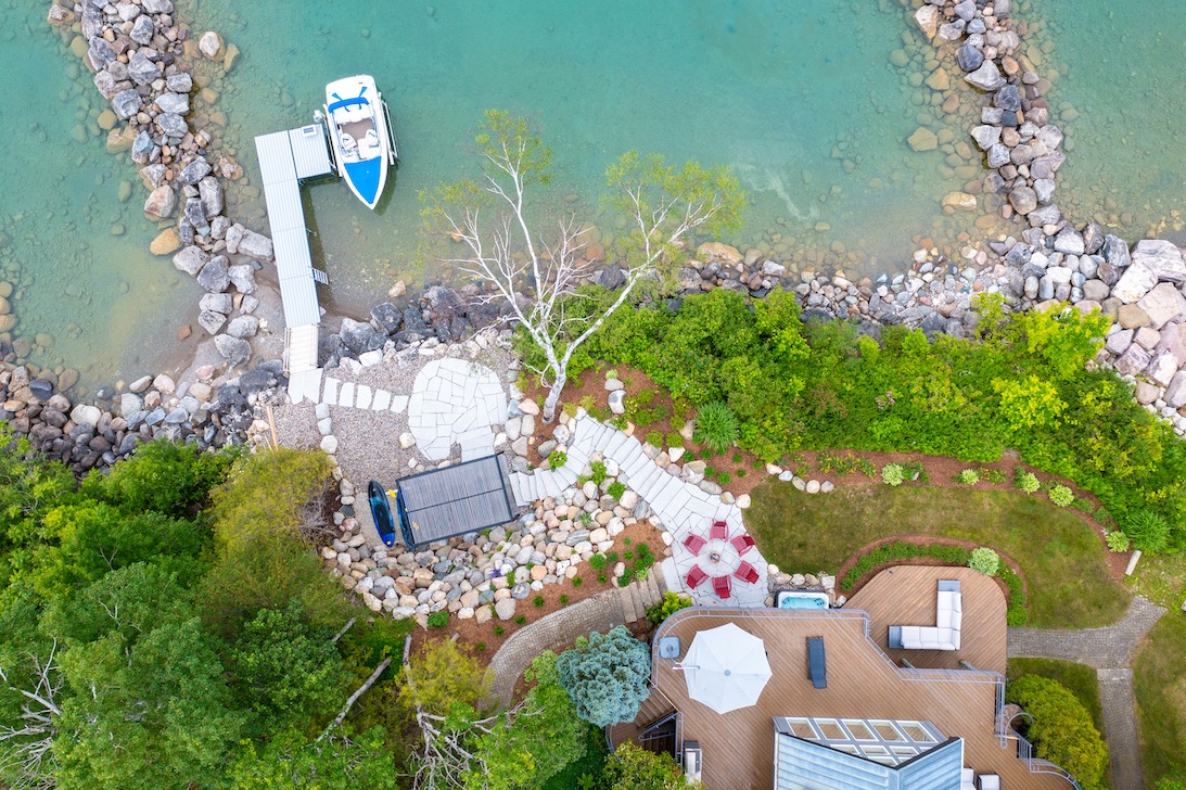 Aerial view of a coastal property with a boat docked at a pier, surrounded by clear waters, landscaped gardens, and a house with outdoor furniture.