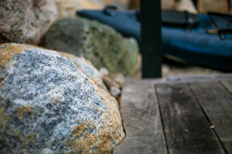 The image shows a close-up of textured rocks in the foreground with a blurry canoe resting on a quiet lakeshore in the background.