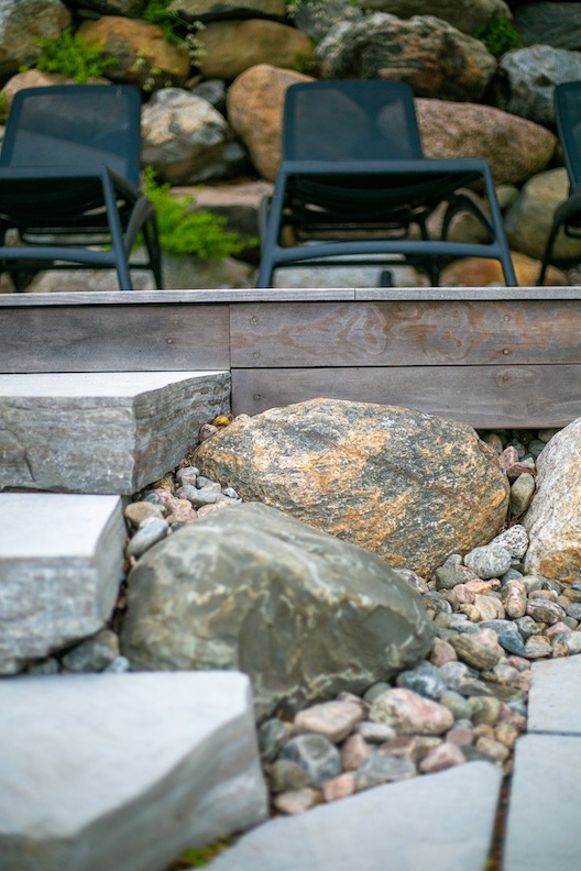 A landscaped outdoor area with a wooden bench, grey stone pavers, large boulders, and smaller pebbles. In the background, there are metal chairs.