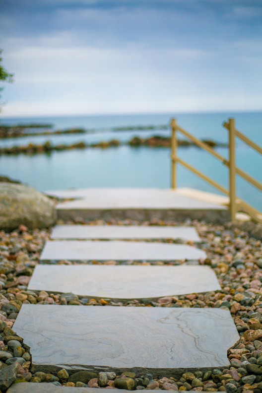 White stepping stones lead down to a tranquil sea, flanked by a wooden railing and pebbled shore, with a soft focus on the serene background.