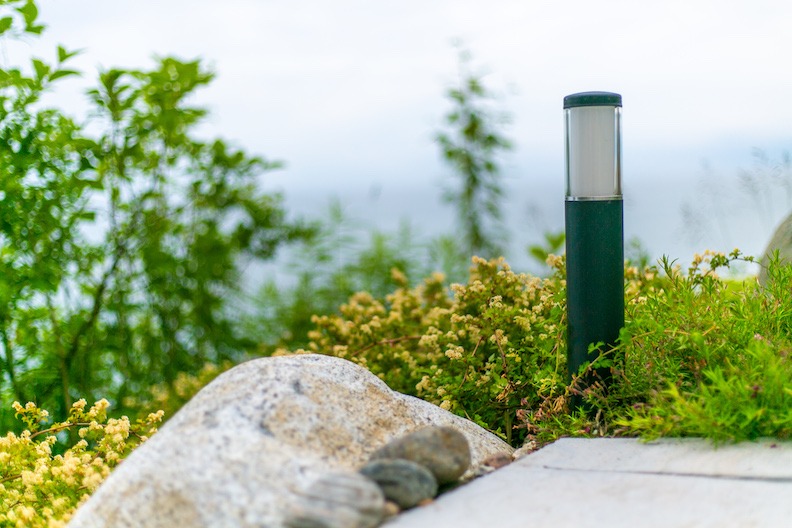 Outdoor setting with a modern bollard light on a pathway, surrounded by lush greenery, rocks, and small yellow flowers, with a hazy background.