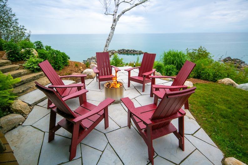 An outdoor patio with red Adirondack chairs arranged around a central fire pit, overlooking a calm sea with a tree, under a cloudy sky.