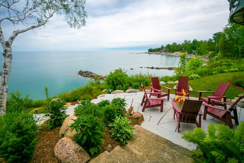 A serene lakeside setting featuring red Adirondack chairs around a fire pit on a stone patio, with lush greenery and a clear view of the calm water.