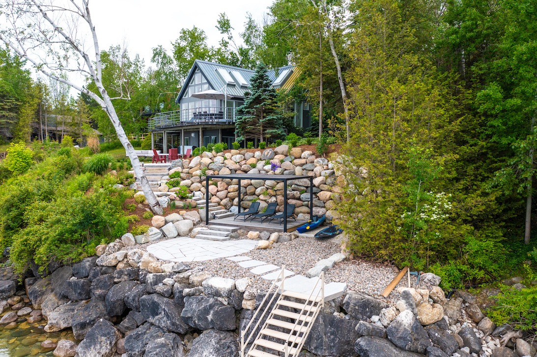 A modern house with large windows stands amidst lush greenery, featuring a landscaped stone path, outdoor seating, and a swing overlooking a rocky waterfront.