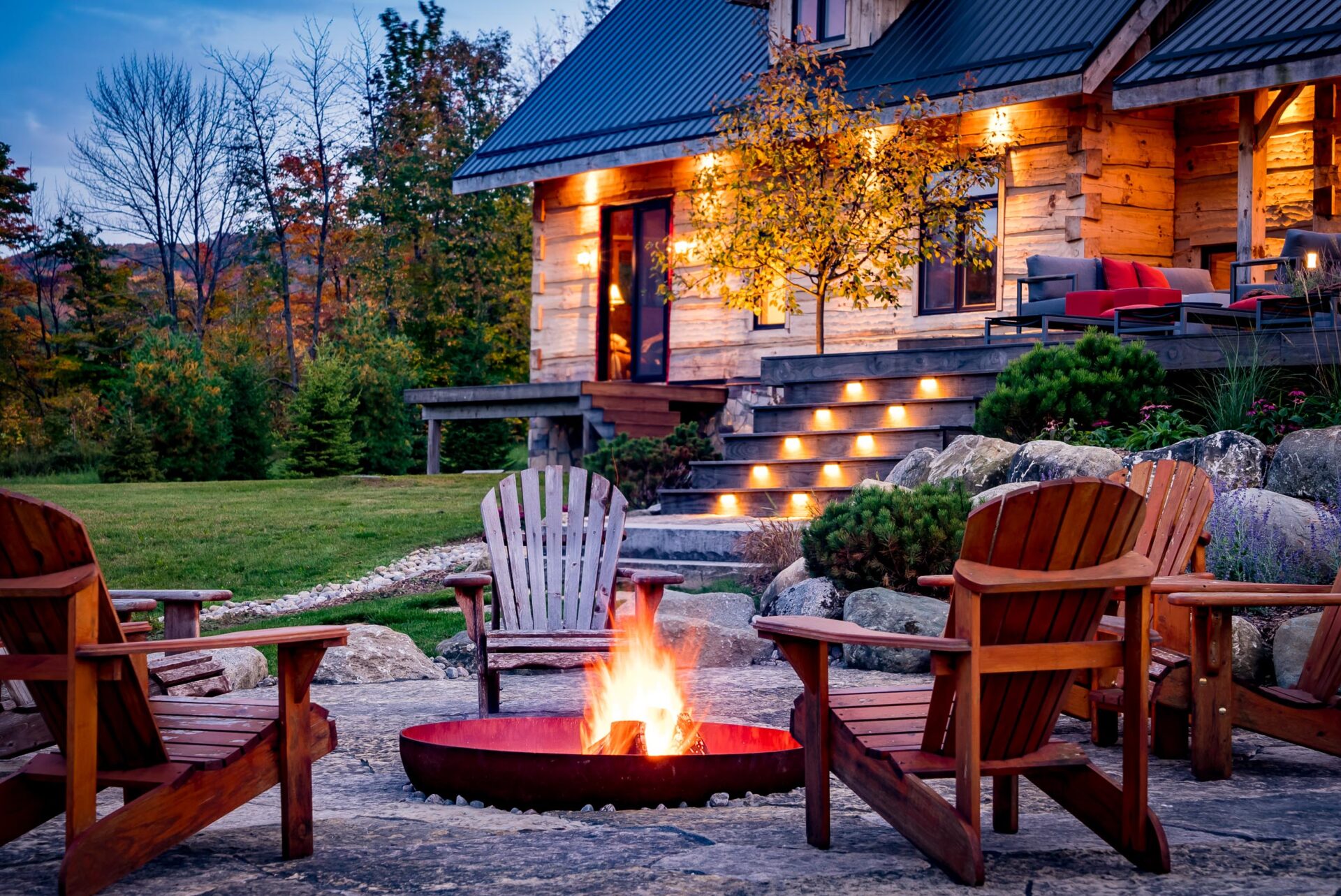 A cozy outdoor fire pit with Adirondack chairs in front of a log cabin during twilight. The steps are illuminated, enhancing the warm, inviting ambiance.
