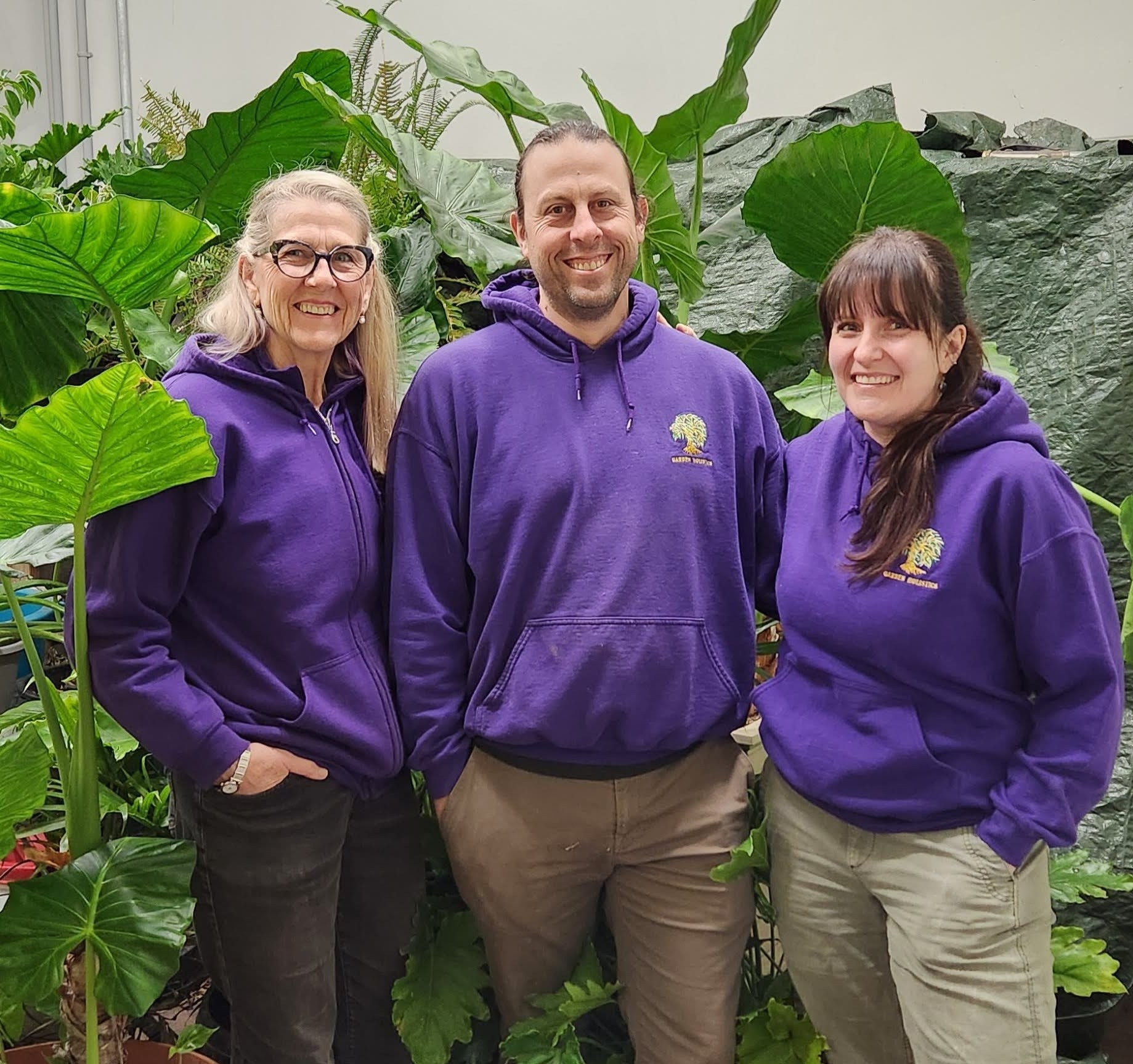 Three people are standing together smiling in a greenhouse, wearing matching purple hoodies with a logo, surrounded by large green leafy plants.