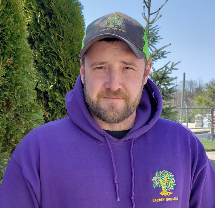 A person wearing a purple hoodie and baseball cap stands outside with trees in the background on a sunny day. They appear contemplative or serious.