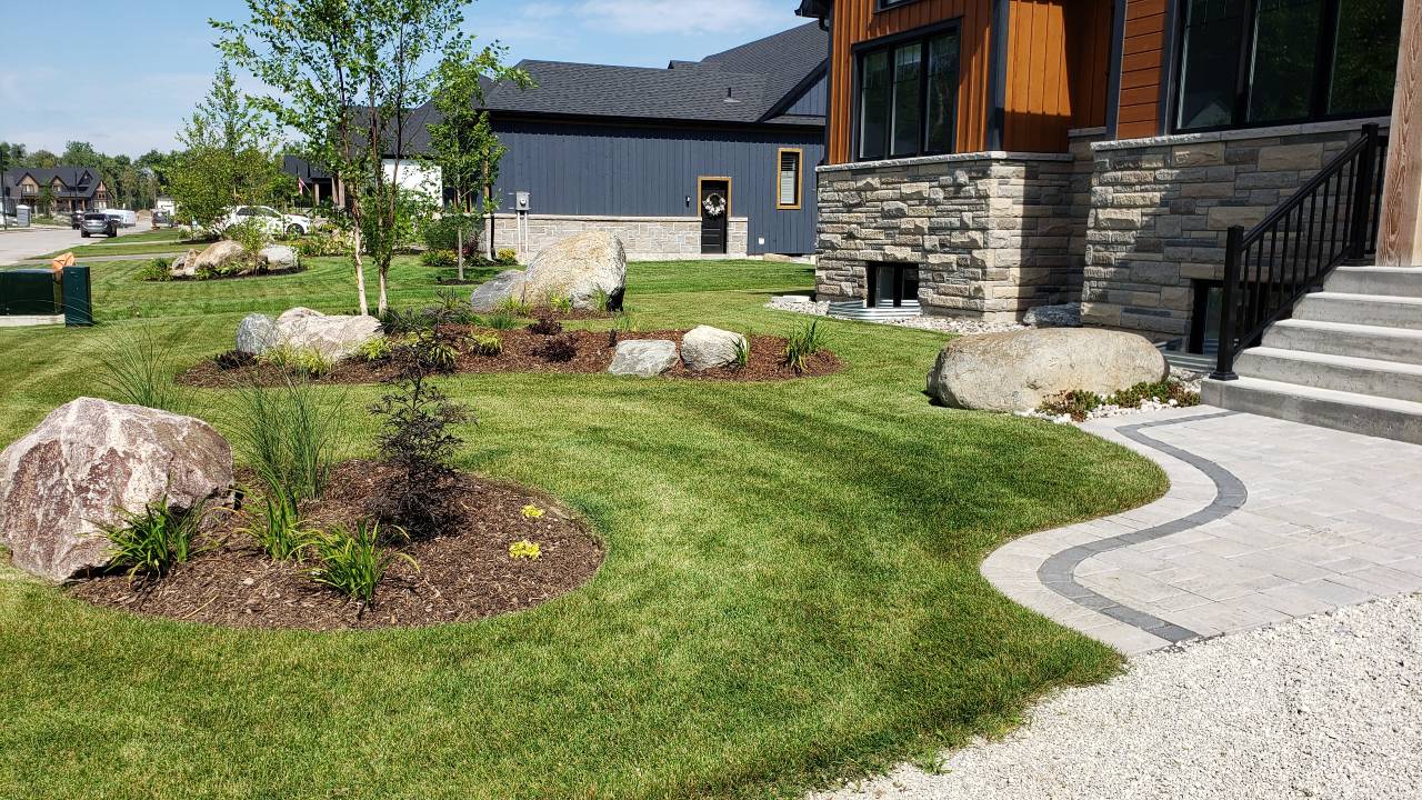 This image shows a landscaped front yard with fresh grass, large stones, plants, and a winding stone pathway leading to a modern house.
