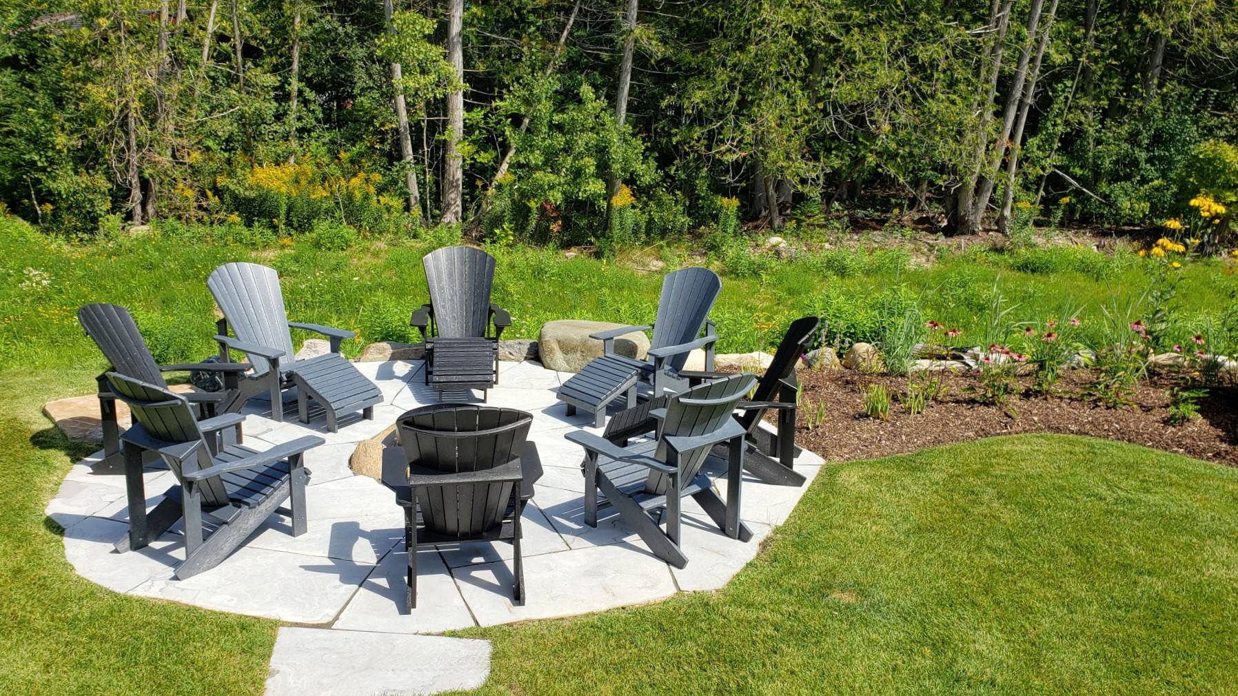 A serene outdoor setting with a circle of grey Adirondack chairs around a fire pit, surrounded by a lush garden and trees on a sunny day.