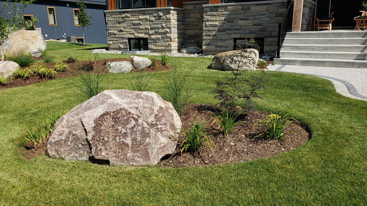 A manicured lawn with decorative rocks, ornamental grasses, a stone house facade, and concrete stairs leading to a porch with seating.