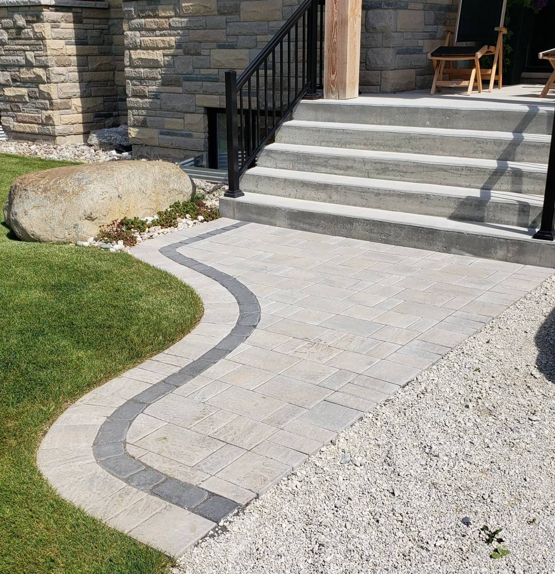 This image showcases a curved paved path leading to a set of concrete stairs with a metal railing, near a well-manicured lawn and stone exterior.