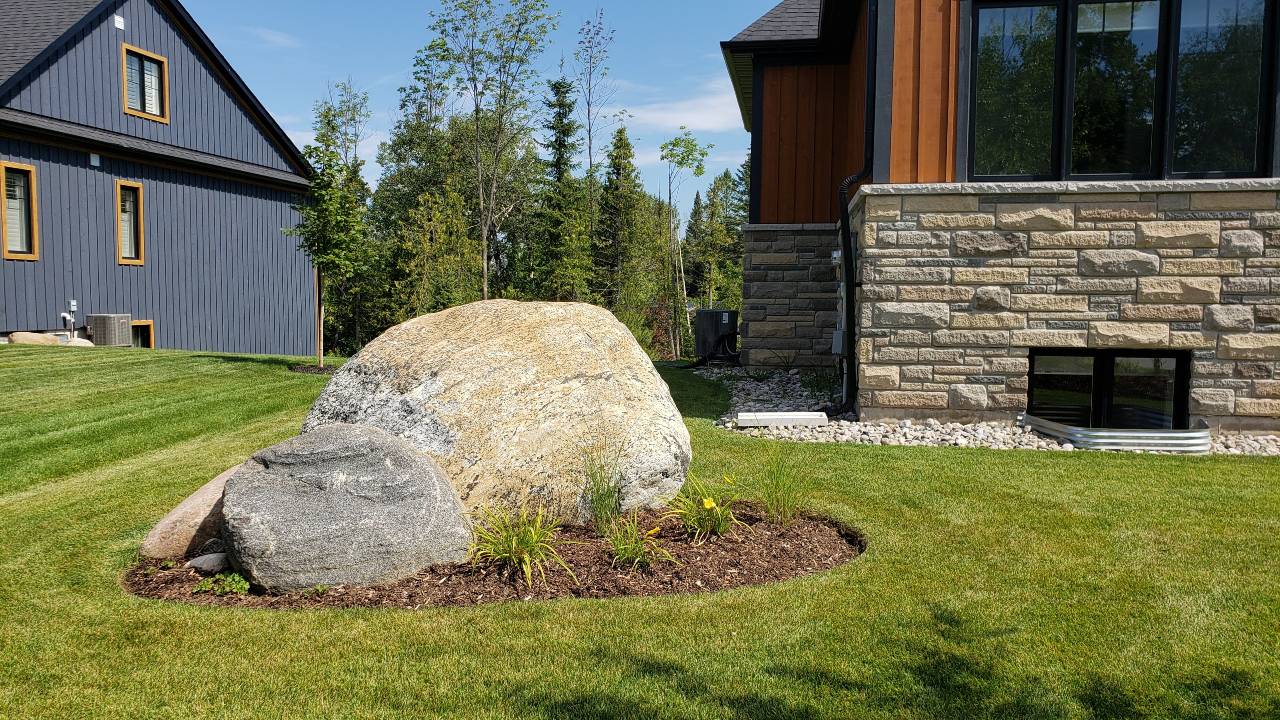 A large rock sits in a landscaped garden bed with young plants, near a modern house with gray siding and stone accents, under a clear sky.