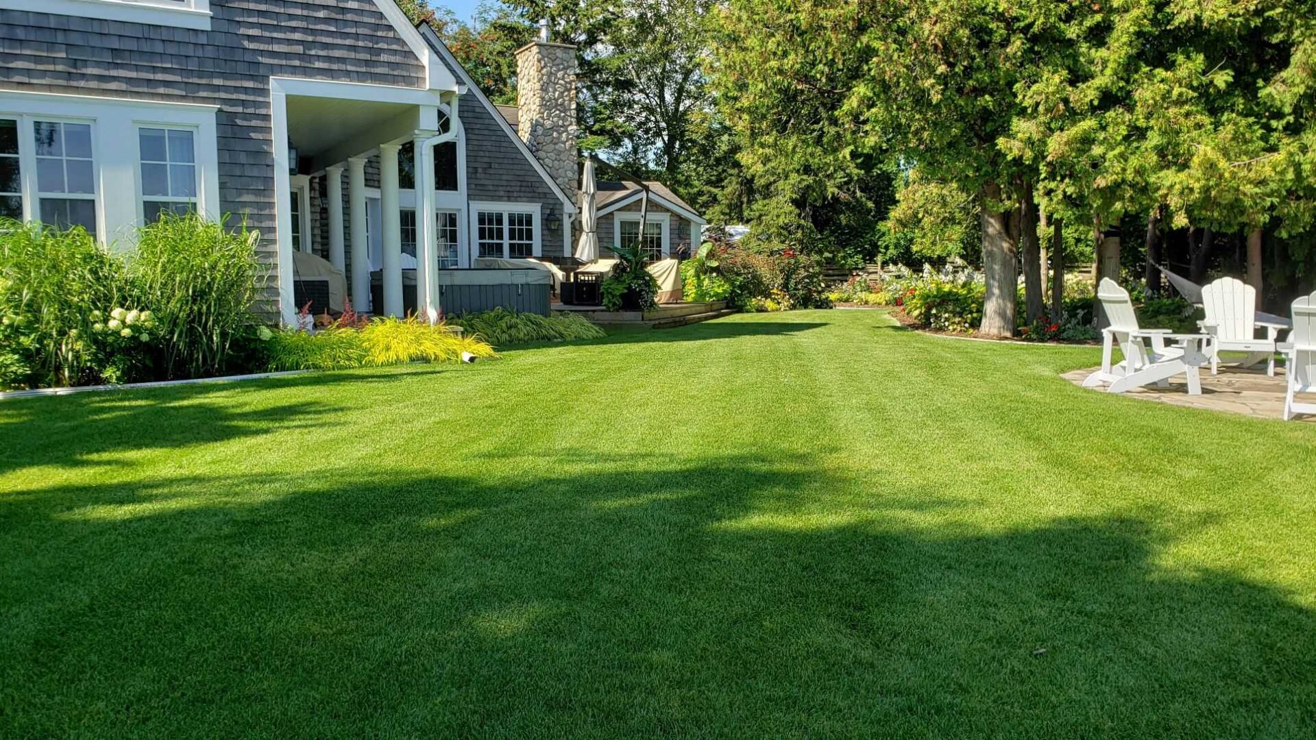 A well-manicured lawn extends before a charming house with a stone chimney, surrounded by lush greenery and white Adirondack chairs in bright sunlight.