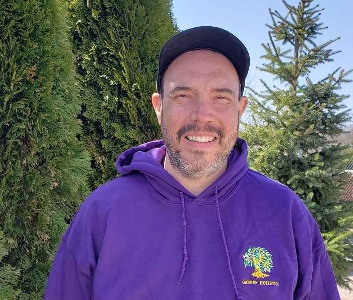 A person wearing a black cap and purple hoodie smiles in front of green conifer trees under a clear blue sky. The sun is shining.